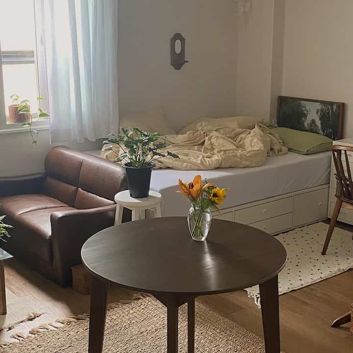 ナチュラルな色調のワンルーム寝室、木製家具とヴィンテージソファ、花瓶のある丸テーブルが配置された雰囲気のあるインテリア