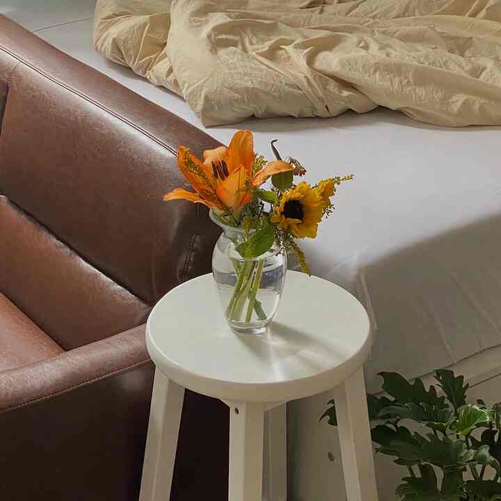 ブラウンレザーのソファとベージュの掛け布団カバーがある寝室、白い丸テーブルの上に花瓶と生花が配置された自然で温かみのあるインテリア