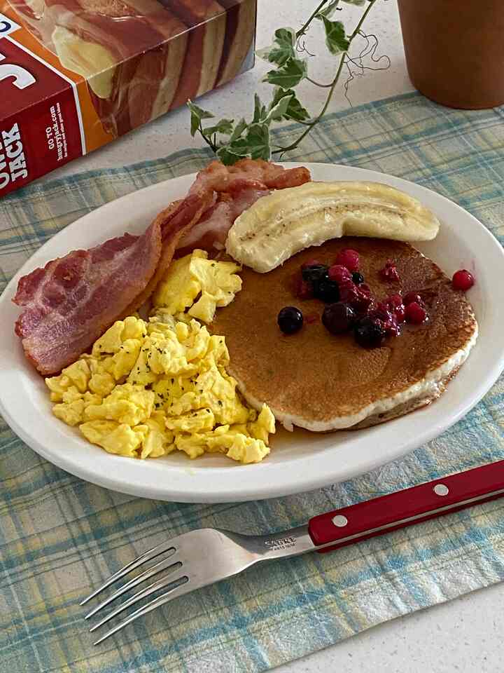Meal setting on a blue and yellow plaid placemat featuring bacon, scrambled eggs, pancake topped with banana and berries