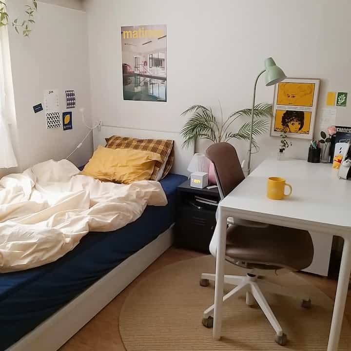 白い壁とネイビーベッドが調和したワンルームの寝室、黄色のアクセントとデスクが配置されたシンプルでナチュラルなインテリア