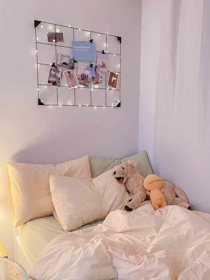 ベージュと白を基調とした寝室、ストリングライトで飾られたメモボードと寝具、犬のぬいぐるみが置かれた温かみのある空間