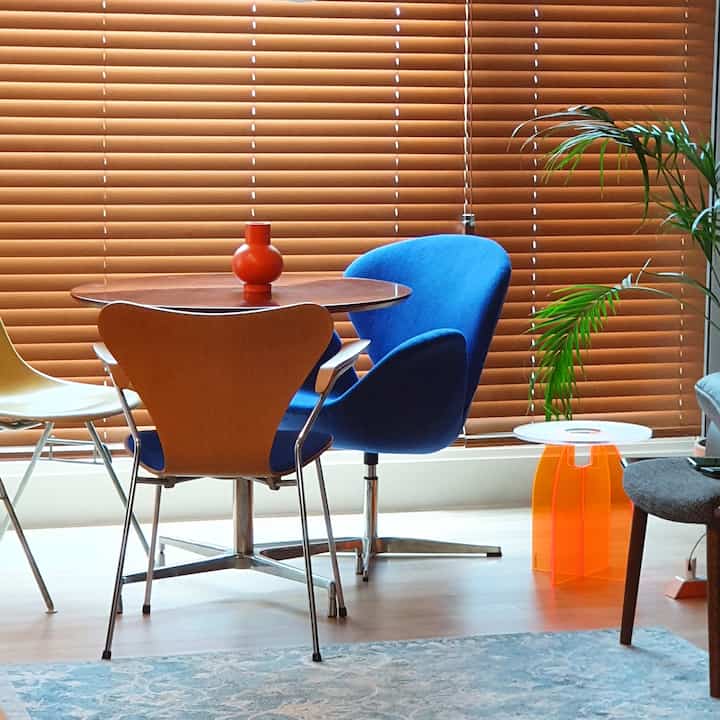 オレンジ色のウッドブラインドを背景にしたリビング、木製の円形テーブルと青いアクリルチェア、オレンジ色の花瓶が配された明るく自然な印象的な空間