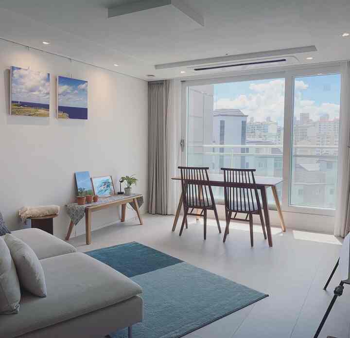 Bright white and natural wood-toned living room featuring a beige sofa, wooden dining table with chairs, and blue rug, creating a cool and airy atmosphere