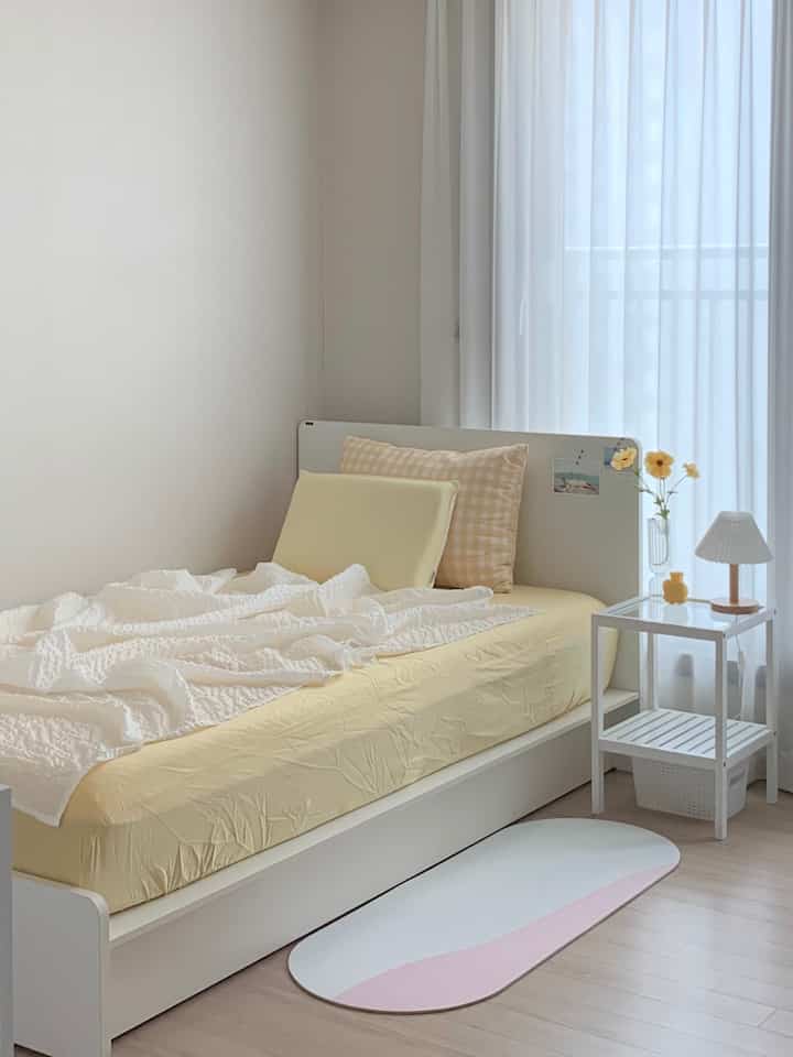 白と黄色の色調の寝室、シングルベッドとナイトテーブル、白いカーテンが配置された温かみのある空間