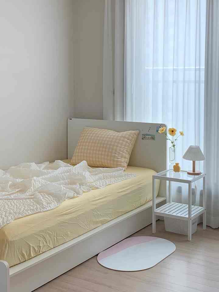 A simple bedroom in white and yellow tones featuring a single bed and side table with lamp, creating a cozy atmosphere