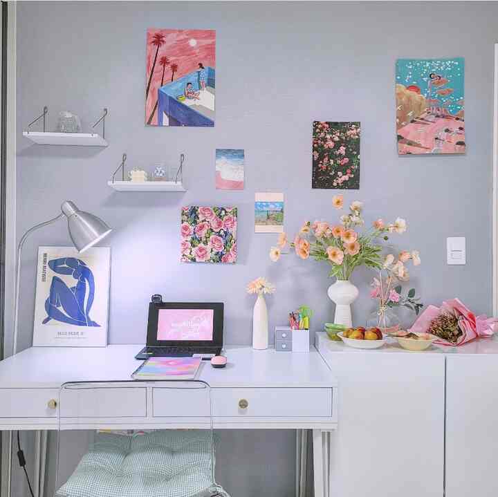 A white and pink toned study featuring a desk, vases with flowers, and various picture frames on the wall creating a cozy workspace