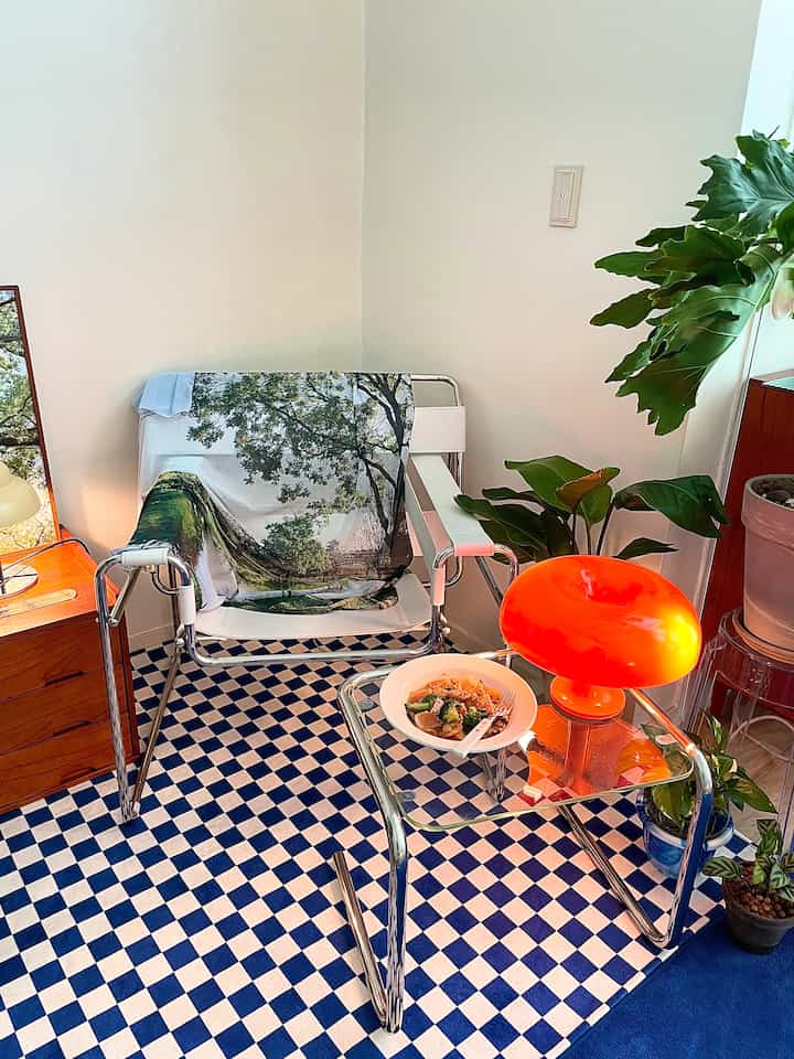 Blue and white checkered rug area featuring chrome metal frame chair, orange table lamp, and green plants, with a modern vibe