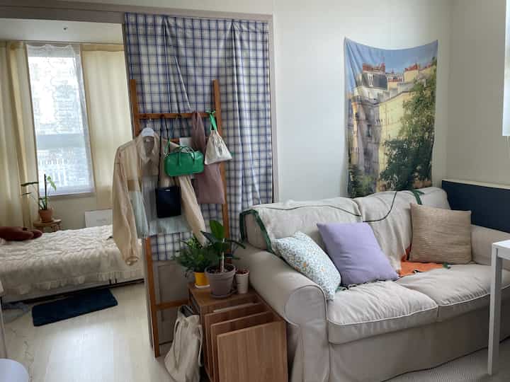 Beige-toned studio living and bedroom space featuring a beige sofa with lavender cushions, wooden coat rack, and plants in a natural setting