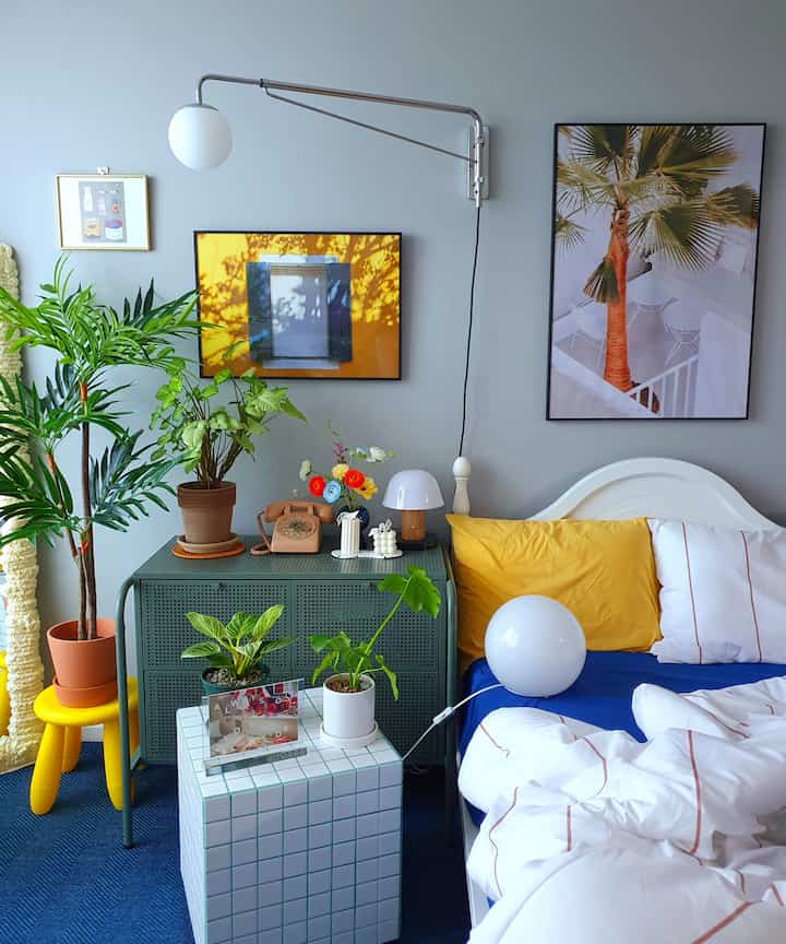 Modern bedroom with white and blue tones, yellow cushions, and abundant plants creating a natural vibe