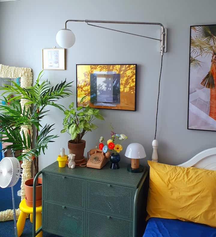 Cozy bedroom with gray walls featuring green dresser, large indoor plants, and yellow pillow with blue bedding