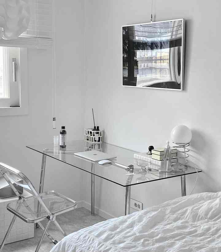 Minimal white-toned bedroom and home office featuring a transparent glass desk and folding chair in a compact single occupancy space