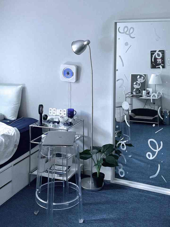 ブルーとネイビーのトーンの寝室、大きな鏡とフロアランプ、透明なバースツールが配置されたモダンで清潔感のある空間