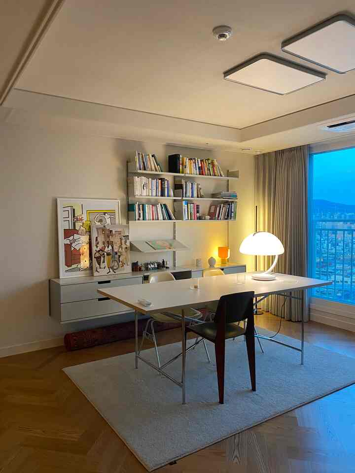 白と茶色のトーンのリビング、金属の棚に本とアートプリント、大きなテーブルと照明が配置された雰囲気のある空間