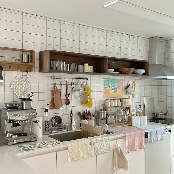 白いタイル壁と木製の棚が調和したキッチン、コーヒーマシンや様々な調理器具が配置された清潔感のある空間