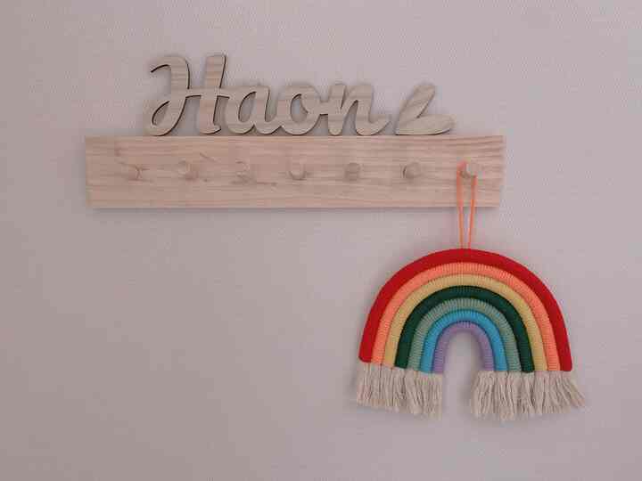 ベージュの壁に木製フックとカラフルなレインボーマクラメが掛けられた、子供部屋の雰囲気ある壁飾り