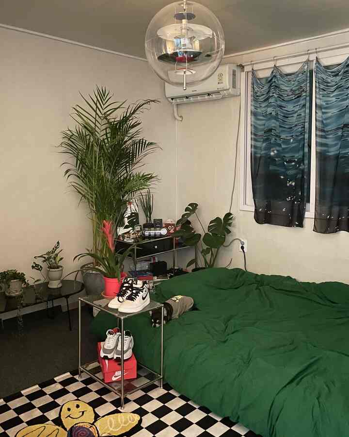 白い壁と天井のワンルーム寝室、緑の寝具と複数の植物、チェッカーボードのラグが配置された雰囲気のあるインテリア