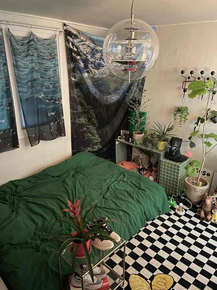 緑の寝具と植物が配置された狭い寝室、チェック柄の床と山の風景ファブリックが印象的な雰囲気のあるインテリア