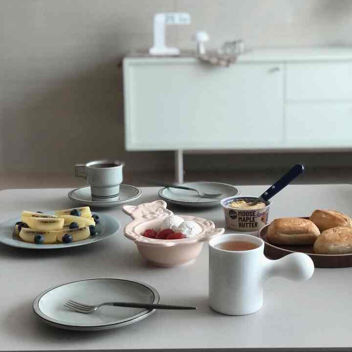 White and gray toned living dining table featuring plates, bowls, and mugs arranged for a simple home cafe breakfast scene