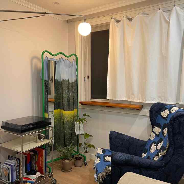 白い壁とカーテン、ブルーのアームチェアがある小さなリビングの一角。緑色の額縁型ハンガーに自然モチーフのファブリックと植物が配置された落ち着いた空間