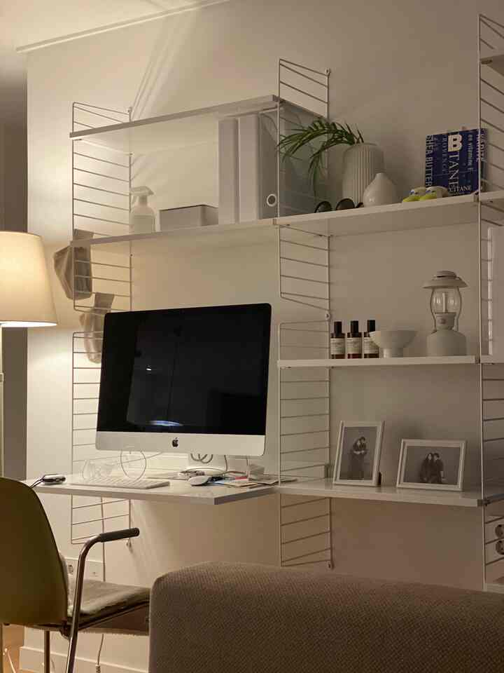 A softly lit white-tone living room home office featuring an iMac, modular shelving system, and a green office chair with a neat aesthetic