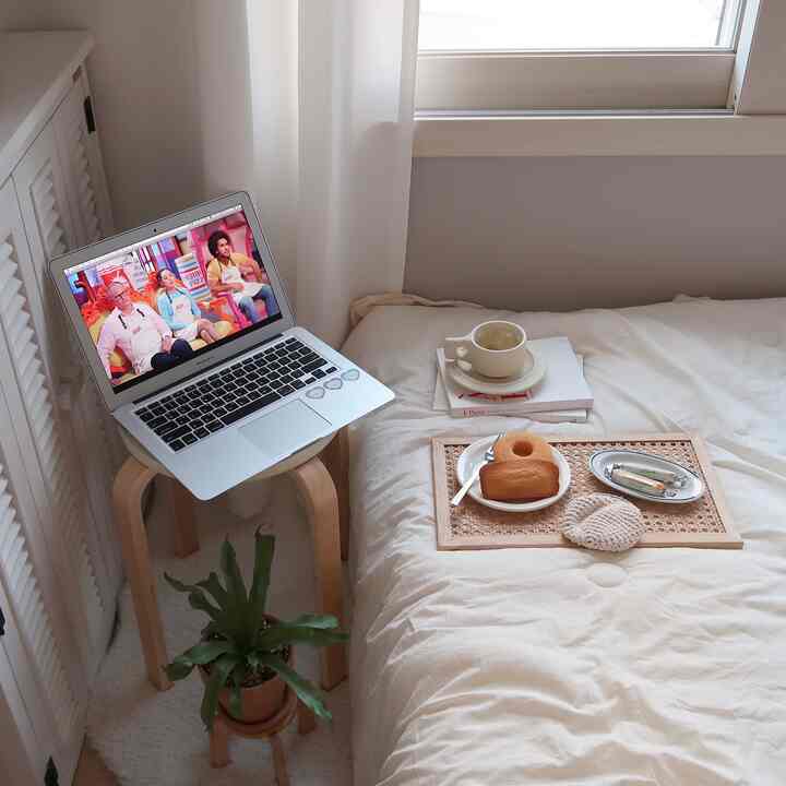 Natural beige bedroom with wooden stool by window, plant pot, and home cafe setup on bed creating cozy atmosphere