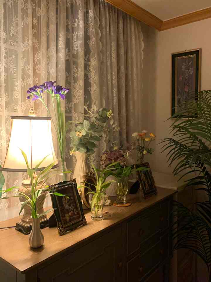 Cozy living room with soft brown wood tones and white lace curtains, featuring various vases and plants on a wooden dresser