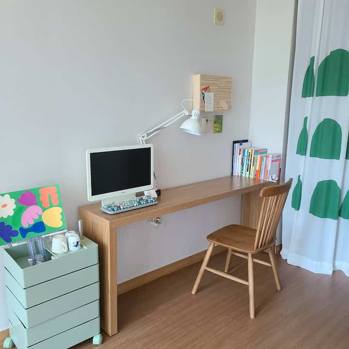 白い壁と木目調の家具が配置されたワンルームのホームオフィス、机の上にコンピューターとテーブルランプが印象的な空間