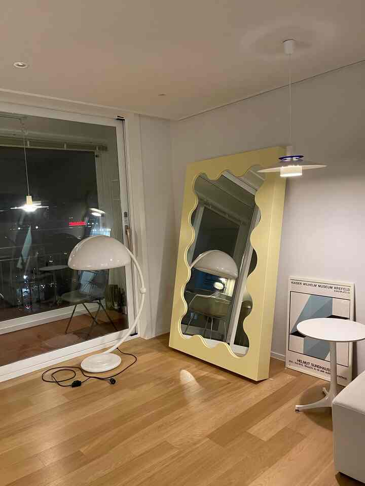 Simple living room with wood tone flooring and pastel yellow framed mirror, featuring white floor lamp and pendant light creating warm atmosphere