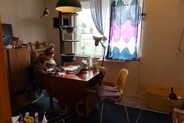 ナチュラルトーンのワークスペース、キャットタワーとデスク、窓際のカラフルなカーテンとフロアランプが配置された印象的な空間