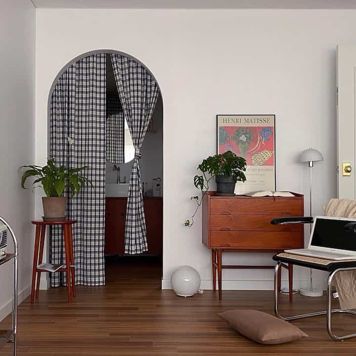 A simple and cozy bedroom with white walls and wood-tone furniture featuring an arched doorway with patterned curtains, console table, and plants