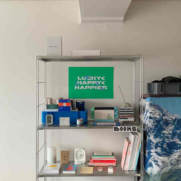 白い壁とメタルシェルフのあるワンルーム、書棚やポスター、時計が配置された印象的な空間