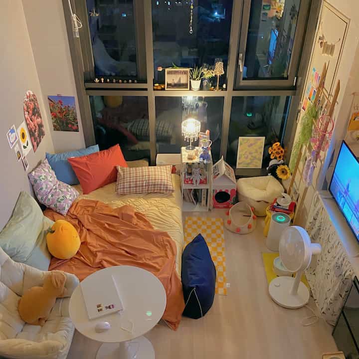 A compact bedroom-living room space dominated by beige and orange tones, featuring a large bed, round table, and cozy window lighting with decorations