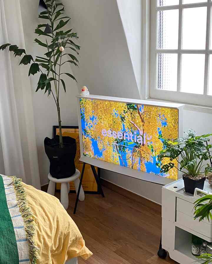 Natural modern bedroom-living space in white and yellow tones featuring a Samsung TV, various plants, stool, and storage cart