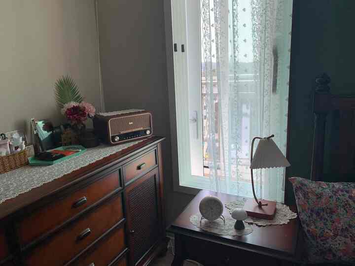 ベージュのトーンの寝室、白いレースカーテンと木製のチェスト、小さなテーブルランプ、かわいらしい小物が配置された落ち着いた空間