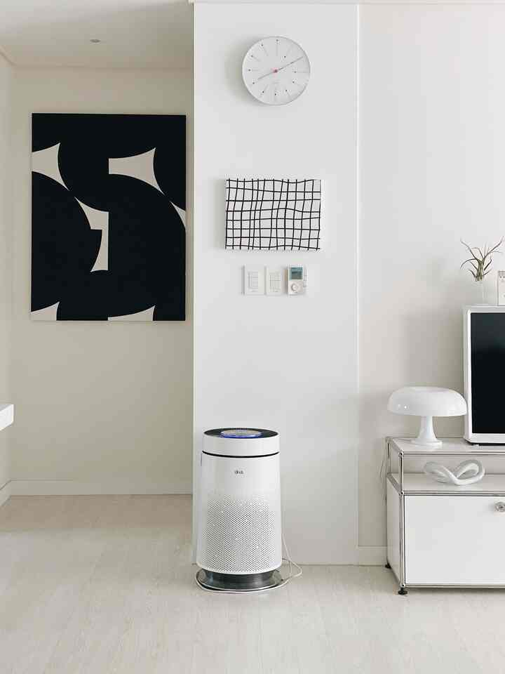 Minimal and modern small living room with white and natural tones, featuring a centrally placed air purifier, wall clock, and table lamp on right
