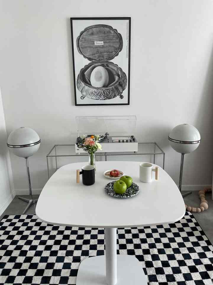 A white-walled living room with a black and white checkerboard rug, featuring a white round square table centered with audio equipment on a transparent rack behind it