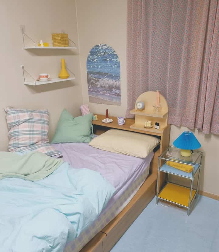 Pastel-toned bedroom featuring lavender bedding, natural wood bed frame, and yellow and mint accents with floral curtains creating a cozy atmosphere