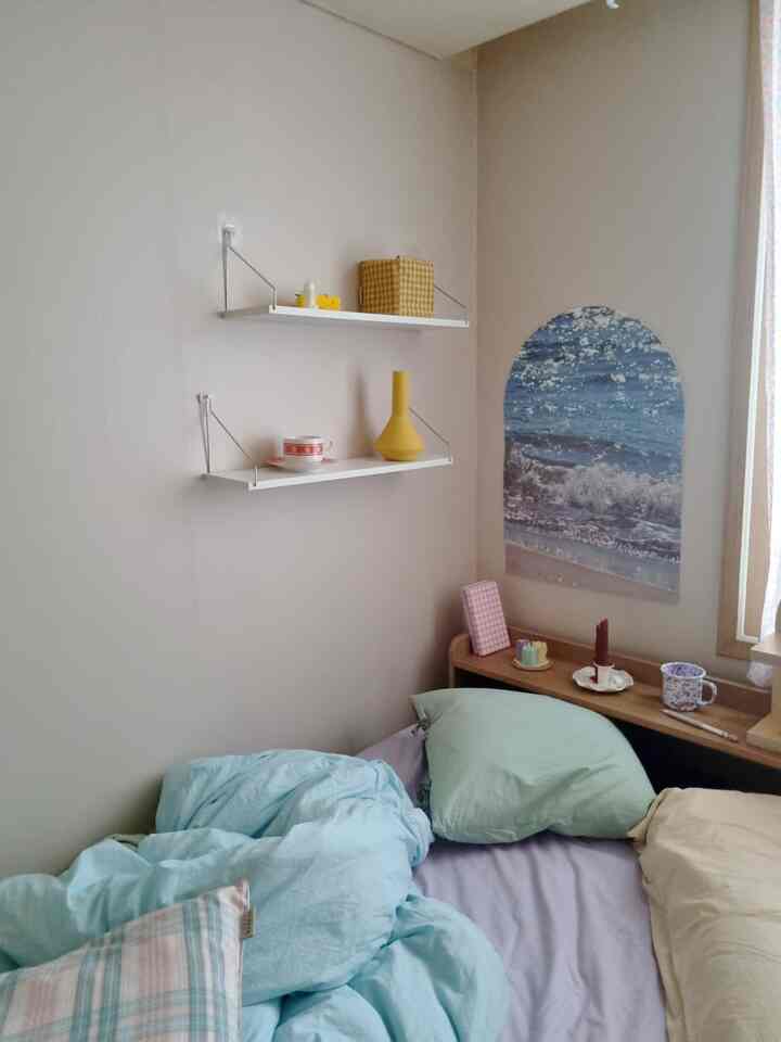A bedroom in soft natural colors featuring a bed with pillows, white floating shelves, and a sea wave poster, creating a cozy atmosphere
