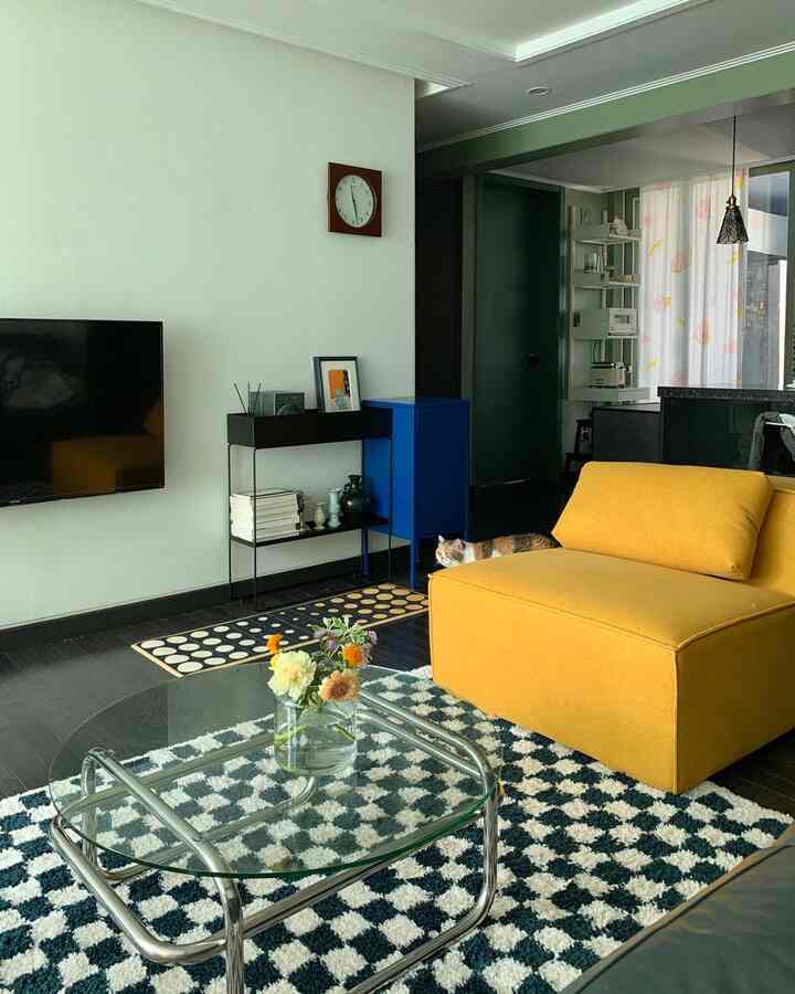 Living room with natural tone walls and dark flooring featuring a yellow sofa, checkerboard rug, and chrome-frame glass coffee table