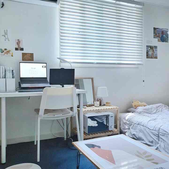 Bright white-toned studio apartment featuring a desk with laptop, single bed with checkered bedding, and small wooden shelving with a microwave, creating a simple personal space