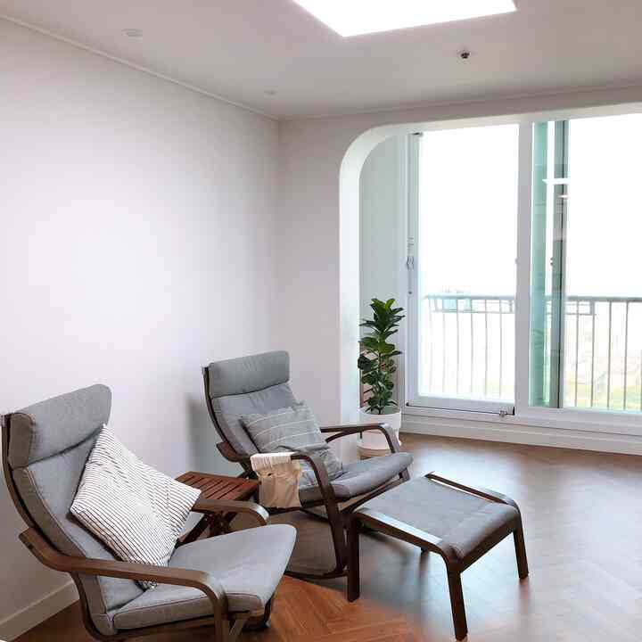 Living room with white walls and brown wooden floor, featuring two gray POÄNG armchairs, a footstool, a plant, and a large window in a simple natural setting
