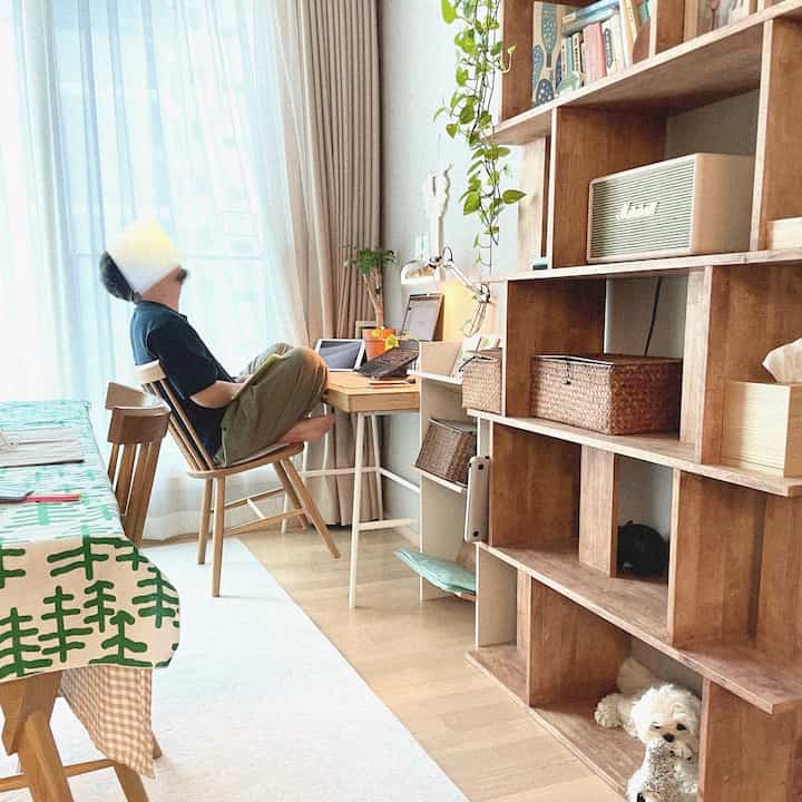 Natural wood tone living room home office featuring wooden dining table, bookshelf, white rug and curtains, with a dog resting under the shelf in a cozy atmosphere