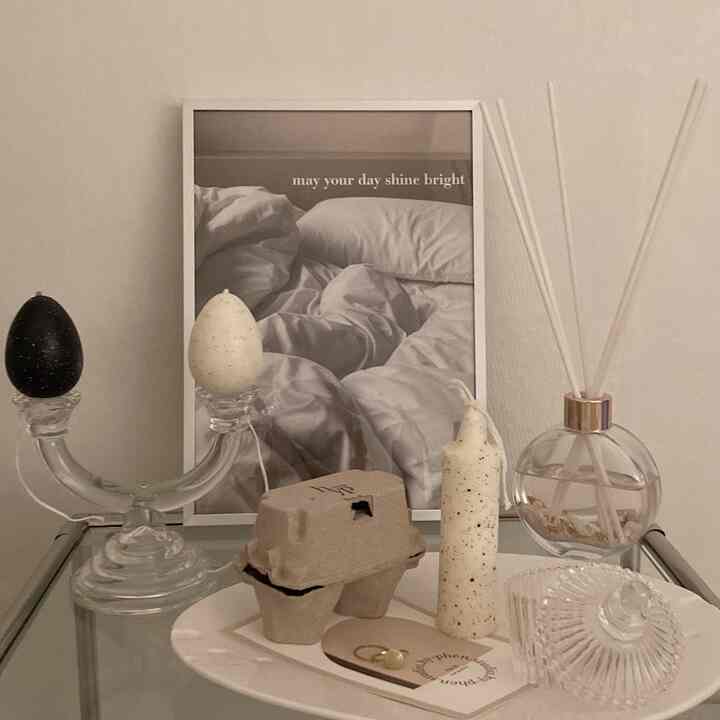 Natural-toned space featuring a transparent modular side table, assorted candles, a diffuser, and a central framed poster against an ivory beige wall, offering a simple and elegant vibe