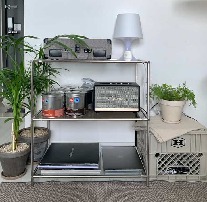 白い壁を背景にしたモジュール棚とミルクボックス、音響機器やコーヒーカプセル、植物が配置された整頓された収納スペース