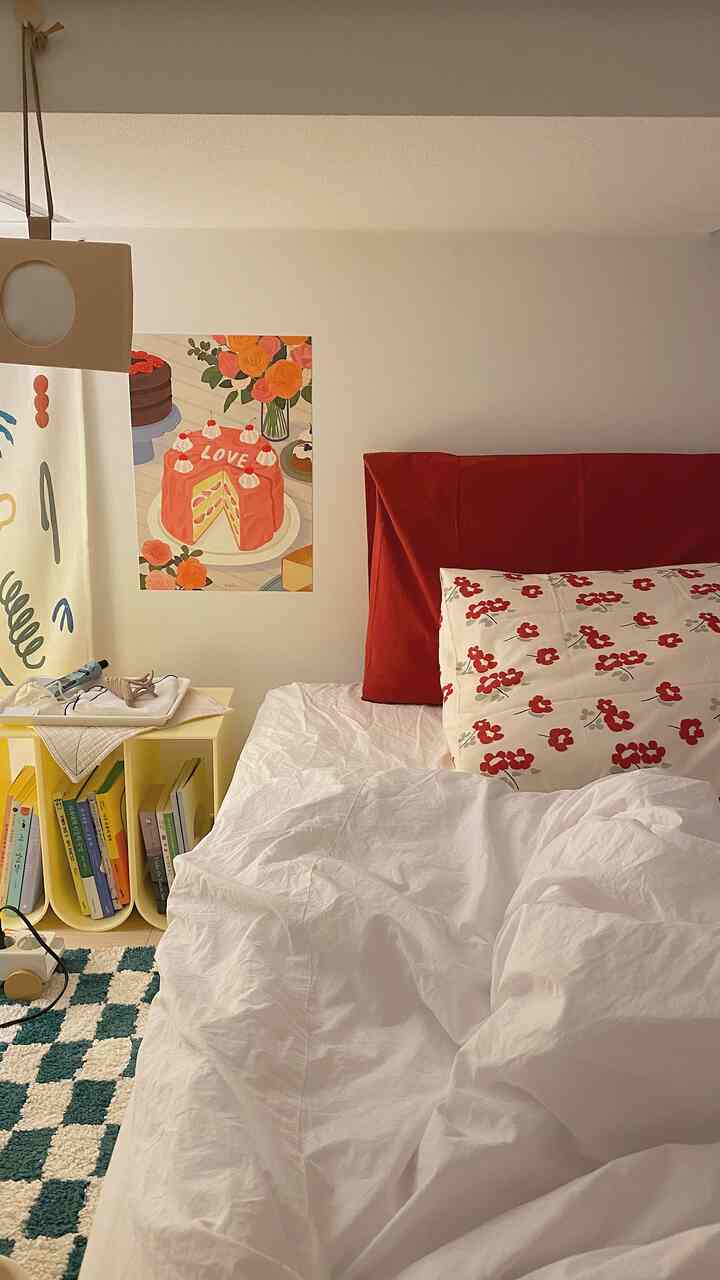A bedroom with white and red tones featuring a red floral pillow, yellow nightstand, and a cake-themed poster, creating a neat atmosphere