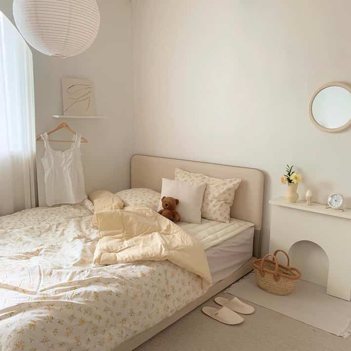 黄色とナチュラルカラーの寝室、花柄の寝具のベッド、暖炉風オブジェ、丸い壁掛け鏡が配置された雰囲気のあるインテリア