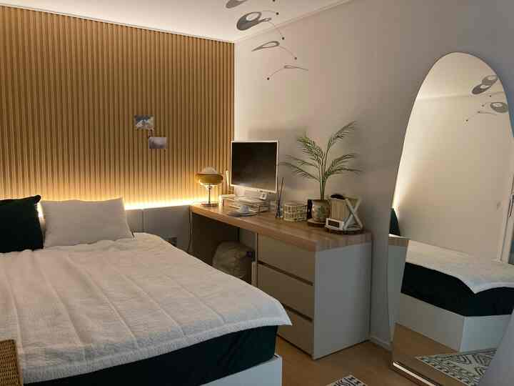 ウッドトーンの壁と白い寝具がある寝室、長いデスクと大型の姿見が配置されたシンプルでナチュラルな雰囲気のインテリア