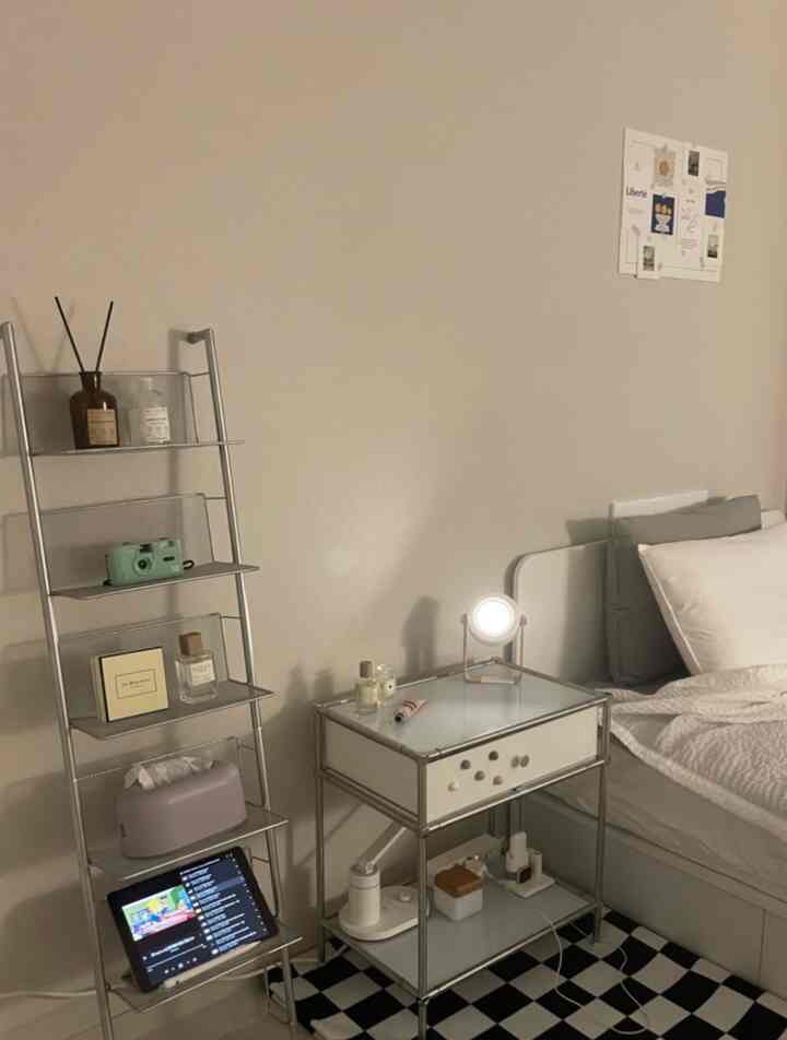 柔らかなアイボリーとホワイトのトーンのワンルーム寝室、ナイトテーブルと棚にディフューザーとアイパッドが置かれ、落ち着いたシンプルな雰囲気の空間