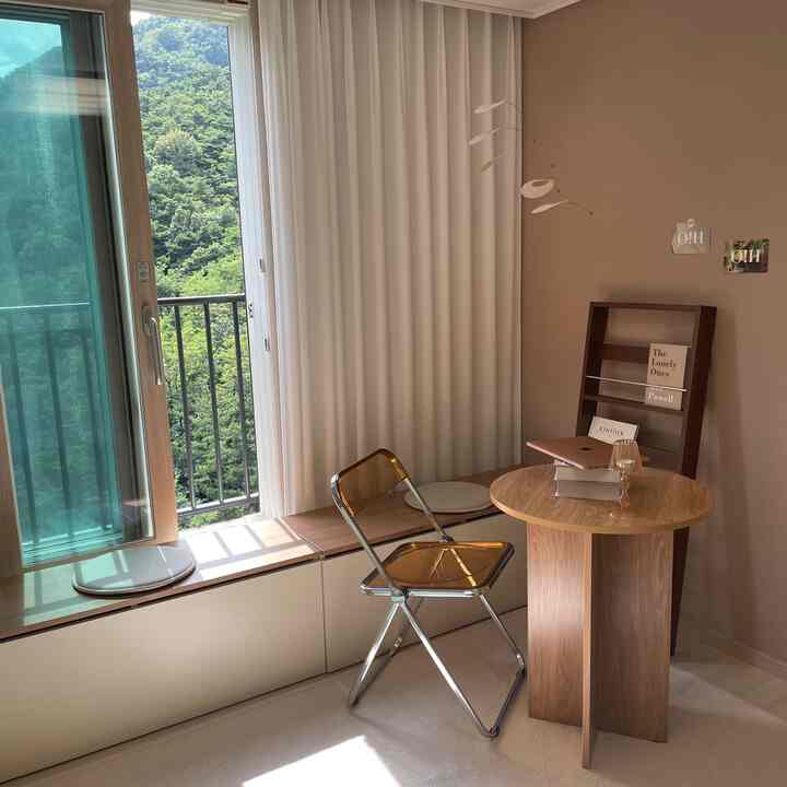Natural brown and white toned study room featuring a round wooden table, folding chair, and long bench storage by the window creating a cozy atmosphere