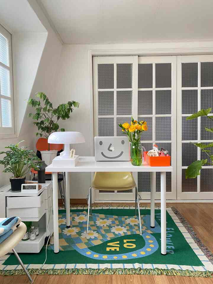 White and green toned bedroom home office featuring white desk, yellow office chair, patterned rug, and multiple plants in cozy setting